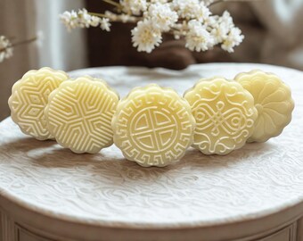 May include: Five round, pale yellow pastries with intricate embossed designs are arranged on a white surface. The pastries are decorated with geometric and floral patterns. White flowers are in the background.