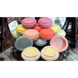 Puede incluir: Colección de bálsamos labiales con forma de macaron de colores, en varios tonos de rosa, naranja, amarillo y azul. Los bálsamos están dispuestos sobre un soporte decorativo de metal negro, algunos están abiertos.