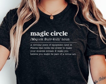 May include: A black t-shirt with white text that reads "magic circle /Maj-ick Surr-kull/ noun A circular piece of equipment used in Pilates that holds the power to make your muscles scream & make you believe you might be part of a circus act."