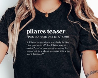 May include: A dark gray t-shirt with white text that reads "pilates teaser /Puh-lah-teez Tee-zur/ noun A Pilates move where your body is like, "Are you serious?" It's Pilates way of saying "you've been doing crunches for years, but how about we make this a lot more dramatic?"