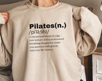 May include: Beige sweatshirt with the word "Pilates" in bold black letters, followed by a phonetic pronunciation and a definition. The definition describes Pilates as a blend of yoga, core work, and breathing exercises.