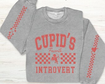 May include: A gray sweatshirt with the text "Cupid's Favorite Introvert" in red. The design includes a cupid with a bow and arrow, and a checkered pattern. Red arrows and hearts decorate the sleeves.