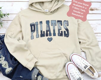 Op de afbeelding: Een beige hoodie met het woord "PILATES" in blauwe letters in patchwork-stijl en een blauw hart. De afbeelding toont ook een blauwe jeans en witte sneakers. De tekst op de afbeelding luidt "Pick your Style! Available in a T-Shirt, Tank, Hoodie or Sweatshirt!"