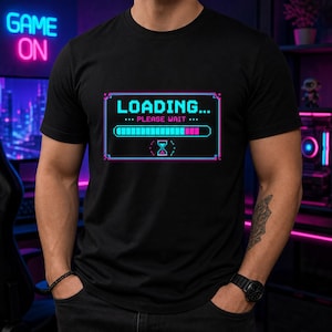 May include: A black short-sleeved t-shirt featuring a central graphic of a pixelated loading bar. The design includes the text "LOADING... PLEASE WAIT" in a cyan font, a progress bar with cyan and pink segments, and a pink hourglass icon.