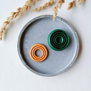 Puede incluir: Dos anillos decorativos, uno naranja y otro verde, sobre una bandeja de hormigón gris. Los anillos están hechos de varios círculos concéntricos.