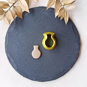 Puede incluir: Un juego de dos cortadores de galletas con forma de jarrón. El cortador más grande es amarillo y el más pequeño es marrón claro. Los cortadores están sobre una superficie de pizarra negra.