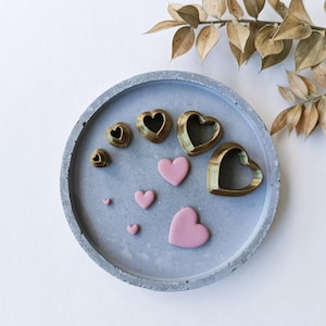 Puede incluir: Un conjunto de moldes en forma de corazón de varios tamaños, hechos de metal dorado, y corazones de arcilla polimérica rosa en una bandeja de concreto gris.