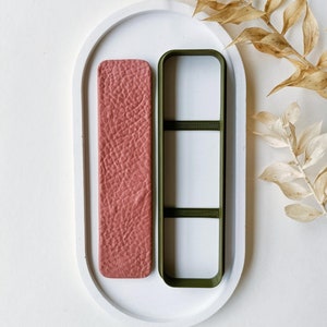 Könnte beinhalten: Zwei rechteckige Silikonformen auf einem weißen ovalen Tablett. Die Form links ist rosa mit einer strukturierten Oberfläche. Die Form rechts ist grün mit einer glatten Oberfläche und drei horizontalen Linien.