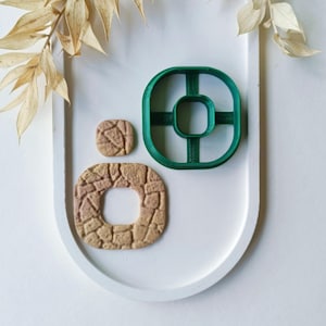 Puede incluir: Un cortador de galletas de plástico verde con un marco cuadrado y un recorte central cuadrado, acompañado de dos piezas de arcilla color canela texturizadas con un patrón de piedra. Una pieza es un cuadrado pequeño y la otra un cuadrado grande con un hueco.