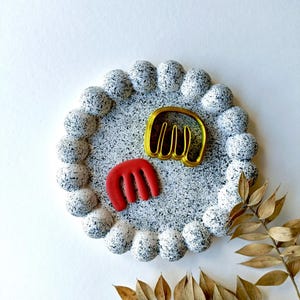 May include: A white ceramic tray with a speckled black and white design and a raised rim. The tray has two small decorative objects on it, one is red and the other is gold.