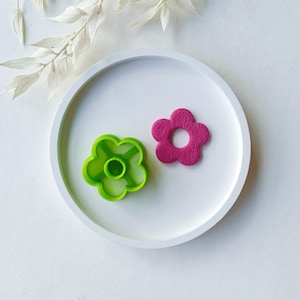 Puede incluir: Dos cortadores de galletas con forma de flor en un plato blanco. Un cortador es verde y el otro es rosa.