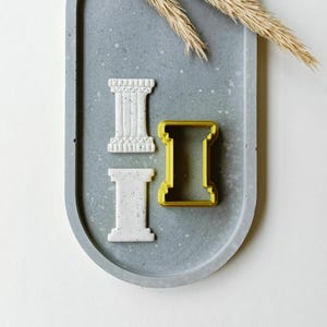 May include: Two cookie cutters in the shape of a column, one white and one gold, on a gray concrete tray.