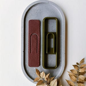 Può includere: Due articoli in pelle di forma rettangolare, uno bordeaux e uno verde oliva, sono esposti su un vassoio ovale in cemento grigio. L'articolo in pelle bordeaux ha un bordo cucito e una forma ritagliata al centro. L'articolo in pelle verde oliva ha un bordo cucito e una forma ritagliata al centro. Il vassoio è su una superficie bianca.
