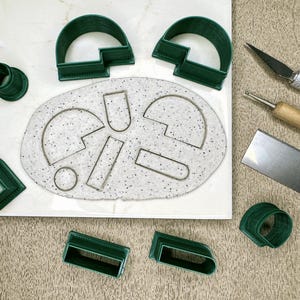 May include: A set of green plastic cookie cutters in various shapes, including circles, arches, and rectangles, arranged on a white surface with a white and gray speckled background. The cutters are used for making cookies or fondant decorations.