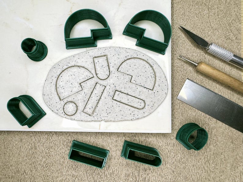May include: A set of green plastic cookie cutters in various shapes, including circles, arches, and rectangles, arranged on a white surface with a white and gray speckled background. The cutters are used for making cookies or fondant decorations.