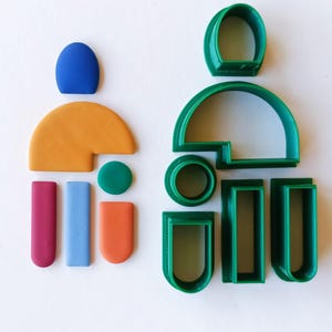 May include: A set of green plastic cookie cutters in various shapes, including a large arch, a circle, a rectangle, and a smaller arch. There are also several small pieces of colorful clay in different shapes.