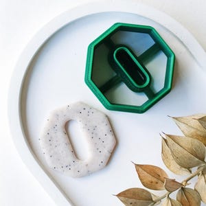 May include: A green plastic cookie cutter shaped like a hexagon with a hole in the center. A white cookie with black sprinkles is next to the cutter.