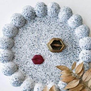 Puede incluir: Una bandeja de cerámica blanca con un diseño moteado en blanco y negro. La bandeja tiene un borde festoneado y presenta un anillo dorado con un diseño de labios rojos en el centro. Un solo amuleto de labios rojos también está en la bandeja.