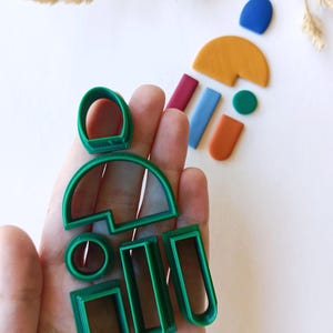 May include: A set of green plastic cookie cutters in various shapes, including circles, rectangles, and arches. The cutters are held in a hand.