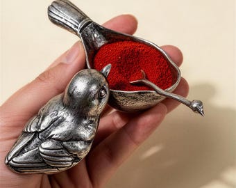 May include: A silver-toned metal bird-shaped container with a textured finish. The container features a hinged lid shaped like a bird and a small scoop resting inside a bowl filled with fine red granules. The metal has a weathered, antique appearance.