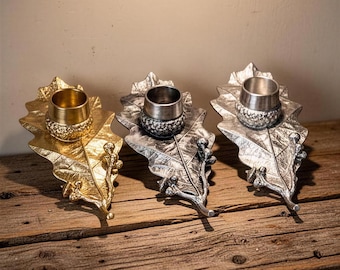 May include: Three metal candle holders shaped like oak leaves with acorn accents. One holder is gold-toned, while the other two are silver-toned with textured surfaces. Each leaf features a central cylindrical cup for holding a single candle.