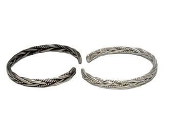 May include: Two silver-toned cuff bracelets featuring a braided, rope-like texture. Each bracelet is an open-ended circle with a metallic finish, showcasing intricate twisted metal strands that create a continuous woven pattern throughout the band.