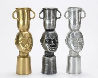 May include: Three decorative vases in gold, dark gray, and silver finishes. Each vase features a sculpted human head as the central body, a cylindrical base, and a top section shaped like a handled vessel with a wide rim and textured details.