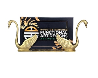 May include: A gold-toned metal business card holder featuring two swan figures facing outward with wings extended. A rectangular black card is held between the swans, displaying the text HUGO DE SÁNCHEZ FUNCTIONAL ART DESIGNS ART WITH.