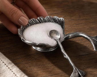 May include: A silver-toned metal salt cellar shaped like a ginkgo leaf with a matching small spoon. The bowl of the cellar is filled with white granular salt. The spoon features a textured handle that resembles a ginkgo leaf stem and leaf.
