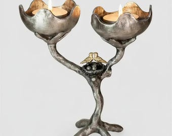 May include: A silver-toned metal candle holder shaped like a tree branch with two flower-shaped cups holding lit tea lights. A small nest with two gold-colored birds sits in the center of the branch structure, supported by a three-legged base.