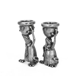 May include: Two pewter frog figurines, each holding a small bowl above their heads. One frog wears a tie, the other a floral adornment. The figures have detailed features and are set against a white background.