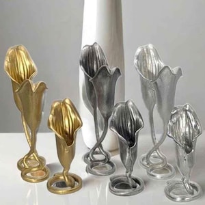 May include: A collection of six metallic flower-shaped vases in gold, silver, and dark grey finishes. Each vase features a flared, petal-like opening and a slender, curved stem that extends down to a circular base, standing in various heights.