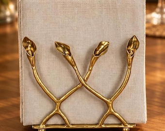 May include: A gold-toned metal napkin holder featuring a base with four small feet and an upright frame shaped like crossed stems topped with four stylized calla lily flower heads, holding a stack of square, light beige textured linen napkins.