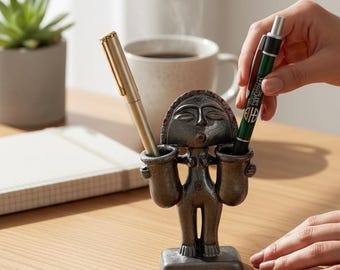 May include: A dark gray, anthropomorphic pen holder featuring a stylized figure with a circular head, small red accents, and two cylindrical containers on either side for holding pens. The figure stands on a rectangular base on a wooden surface.