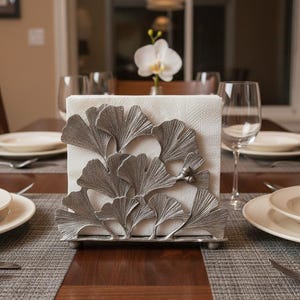Op de afbeelding: Een zilverkleurige servethouder met een ginkgo bladeren ontwerp op een houten tafel. De houder is gevuld met witte servetten en maakt deel uit van een tafelsetting met borden, bestek en wijnglazen. Een witte orchidee staat op de achtergrond.