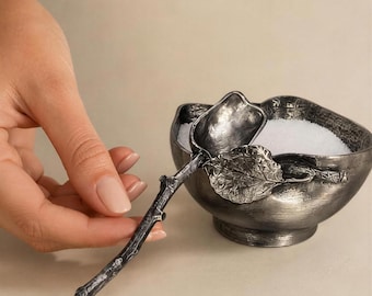 May include: A dark metallic bowl with a textured, weathered finish containing white granules, accompanied by a matching spoon shaped like a tree branch with a prominent leaf detail attached to the handle, featuring an antique silver-toned appearance.
