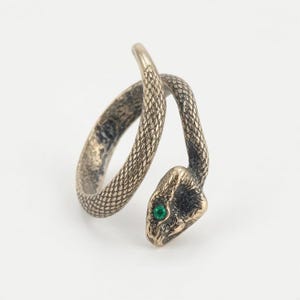 Puede incluir: Un anillo abierto con forma de serpiente enroscada con un acabado en bronce envejecido y texturizado. El cuerpo de la serpiente presenta patrones de escamas detallados a lo largo de toda la banda y la cabeza tiene una pequeña piedra redonda de color verde vibrante.