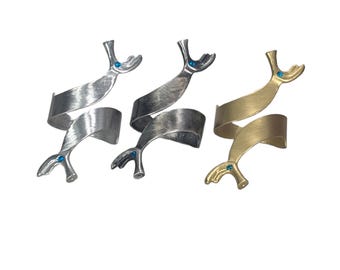 May include: Three spiral-shaped rings, each featuring a whale tail design at both ends. One ring is polished silver, one is brushed dark gray, and one is matte gold. Each whale tail is accented with a small, circular, bright blue gemstone.