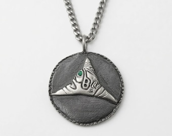 May include: A circular silver pendant with a dark, oxidized finish featuring a raised, abstract triangular relief design and a single small green gemstone accent. The pendant hangs from a matching silver curb chain with a textured edge detail.