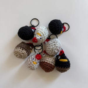 May include: A collection of seven small, round, crocheted chicken keychains in black, white, and brown yarn. Each chicken features a red crocheted comb, a yellow beak, and two small plastic googly eyes attached to a black metal keyring.