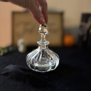 May include: A clear, vintage-style glass perfume bottle with a ribbed body and a faceted stopper. The bottle is held by a hand, showcasing its delicate design. The background is blurred, with hints of other objects.
