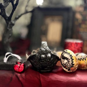 May include: A black decorative bowl with cherub and floral details, alongside a red heart-shaped ornament and two decorated eggs. The eggs have gold and black designs, including a raven and skull. The items are on a red surface.