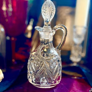 May include: A clear glass decanter with a decorative stopper and handle. A silver crescent moon charm hangs from the neck. The decanter has a textured, cut-glass design. The background includes a red glass and a white candle.