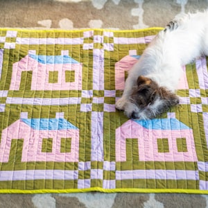 May include: A square quilted blanket featuring a repeating pattern of four pink houses with blue roofs, separated by olive green and light purple checkered borders, with a bright yellow edge binding and a small white and brown dog resting on top.