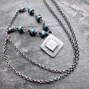 May include: A silver necklace with a diamond-shaped pendant and turquoise beads. The pendant has a textured surface with a smaller square in the center. The necklace chain is a delicate link style.
