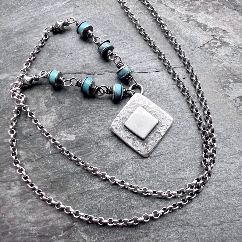 May include: A silver necklace with a diamond-shaped pendant and turquoise beads. The pendant has a textured surface with a smaller square in the center. The necklace chain is a delicate link style.