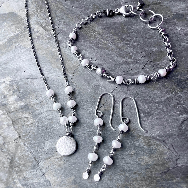 May include: A jewelry set featuring a necklace, bracelet, and earrings. The necklace has a chain with white pearls and a textured pendant. The bracelet and earrings also feature white pearls. All pieces have a silver-toned metal.