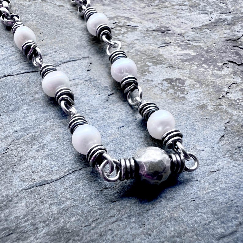 May include: A close-up of a necklace featuring white, round beads and a hammered silver-colored focal bead. The beads are strung on a silver-colored chain with coiled wire accents, creating a delicate and elegant design.