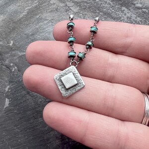 May include: A silver necklace with a diamond-shaped pendant and turquoise beads. The pendant has a textured border and a square center. The necklace is held in a hand, showcasing the intricate design and craftsmanship of the jewelry.