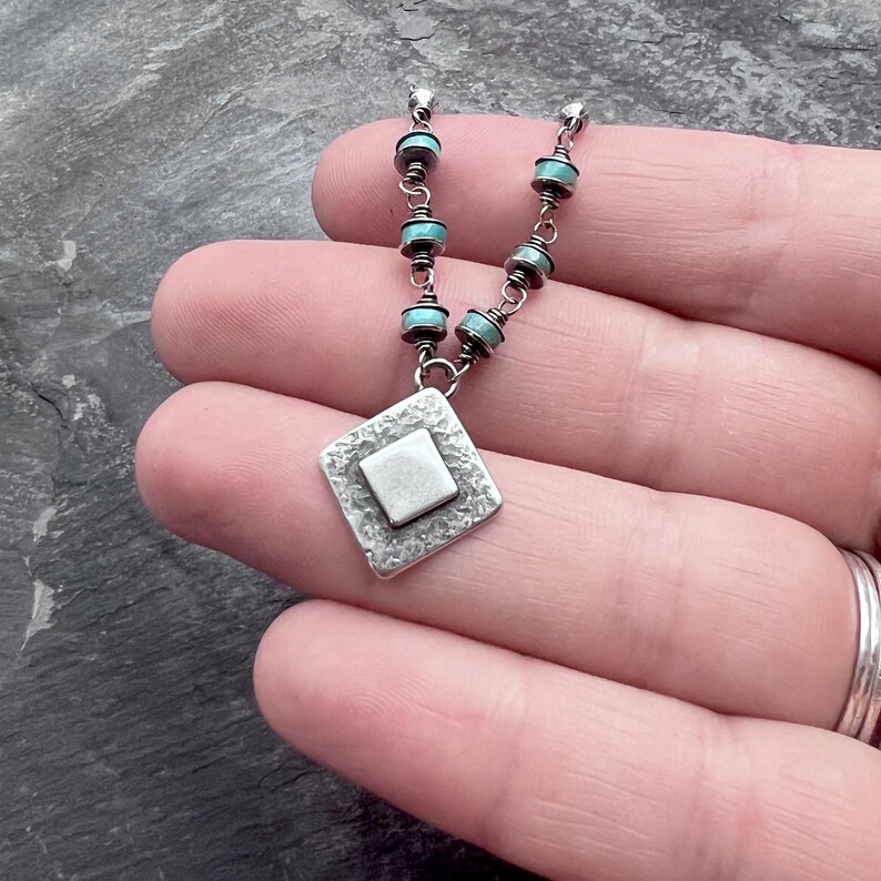 May include: A silver necklace with a diamond-shaped pendant and turquoise beads. The pendant has a textured border and a square center. The necklace is held in a hand, showcasing the intricate design and craftsmanship of the jewelry.